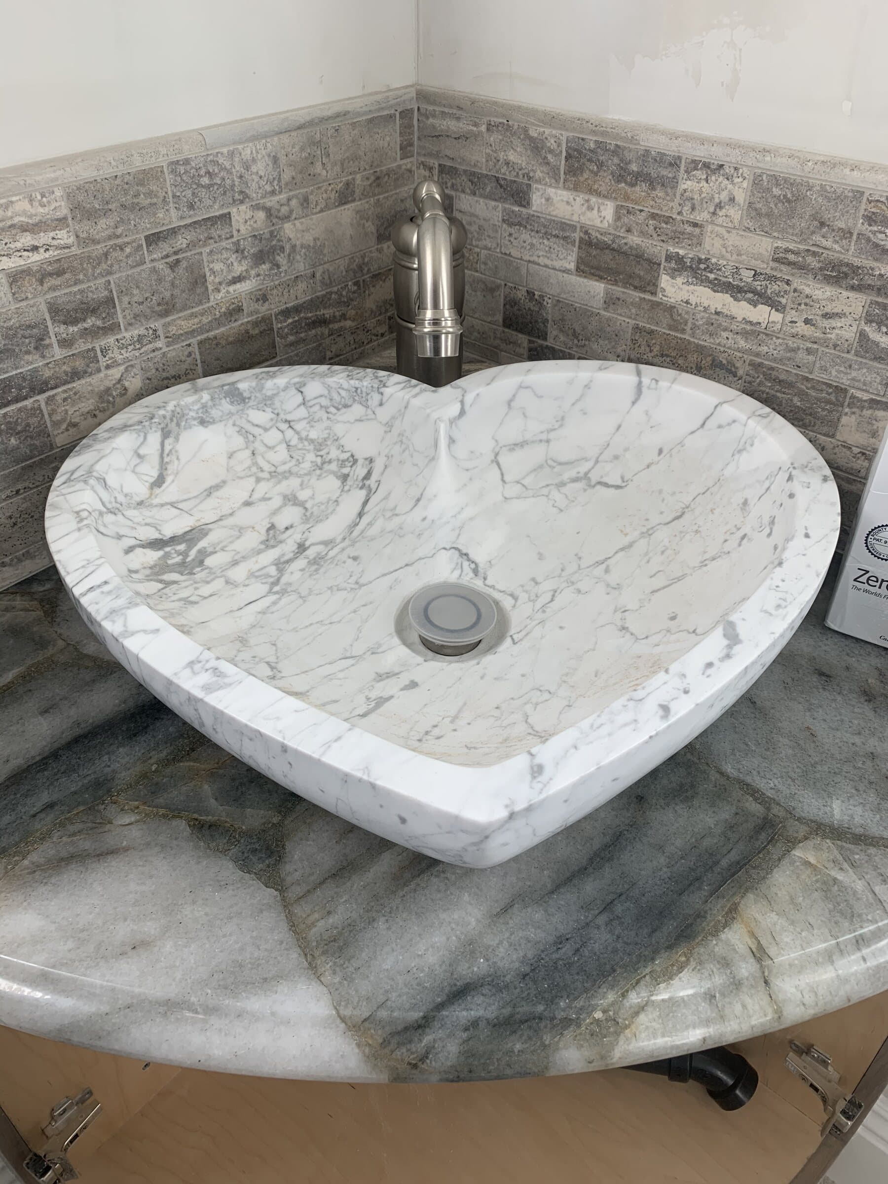 Heart-shaped Carrara marble vessel sink set on a quartzite countertop against a stacked stone tile backsplash