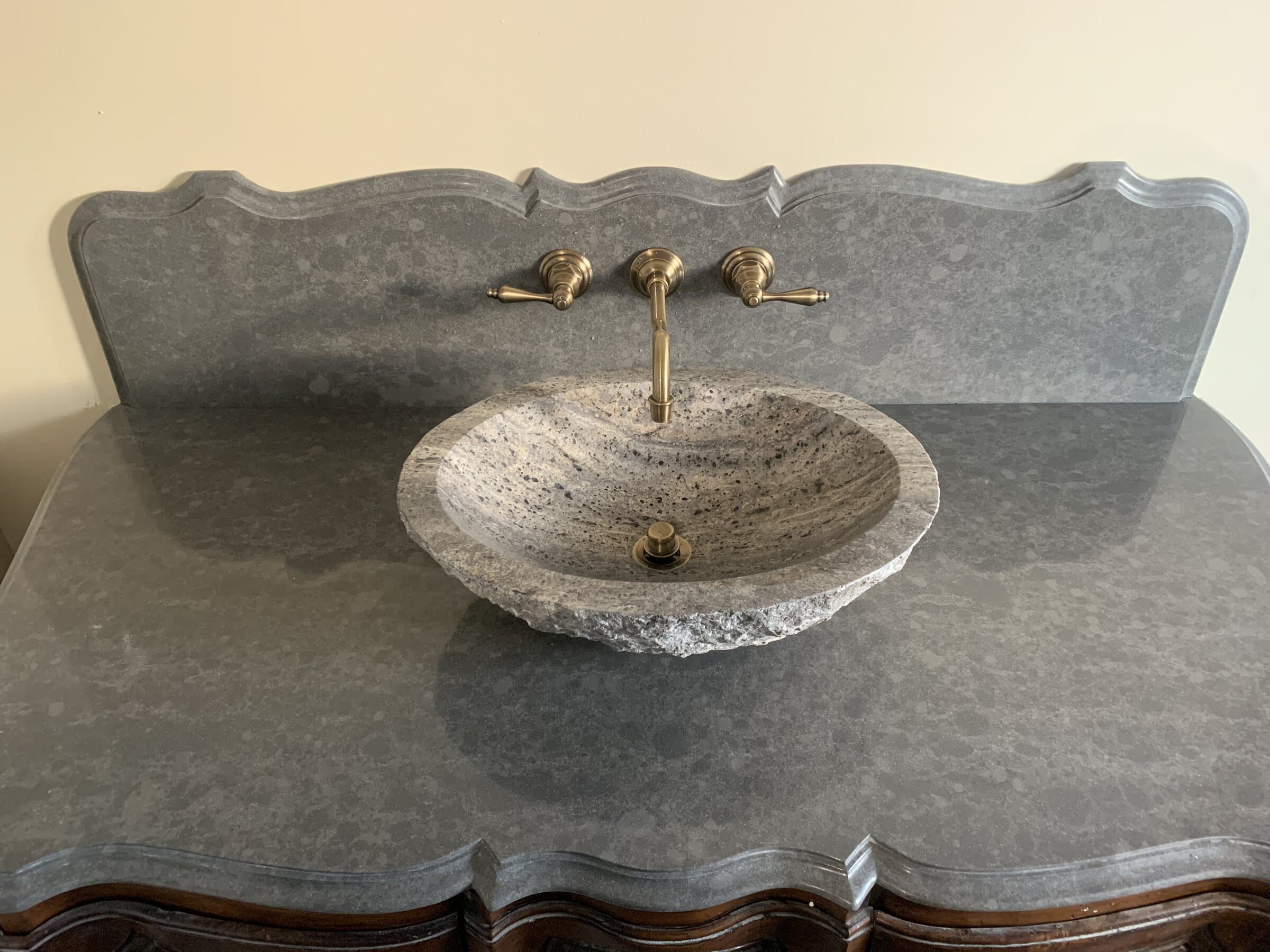 Carved stone vessel basin on a bluestone vanity top with an unlacquered brass wall-mounted widespread faucet and cross handles
